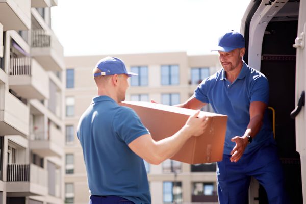 Movers unloading a cardboard box from a van in front of a modern apartment building.