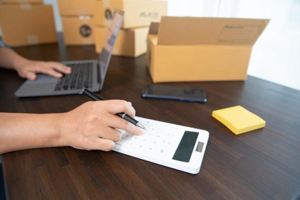 Person using calculator and laptop at desk with cardboard boxes and sticky notes around.