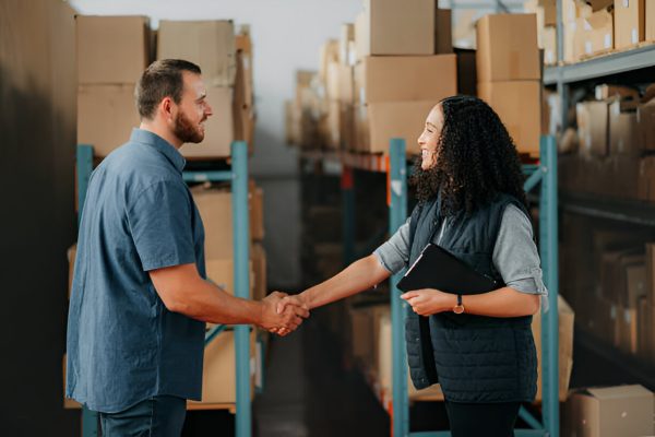Two people shaking hands in a warehouse, confirming a storage or logistics agreement among packed boxes.
