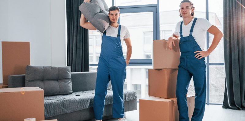 Busy day. Two young movers in blue uniform working indoors in the room.